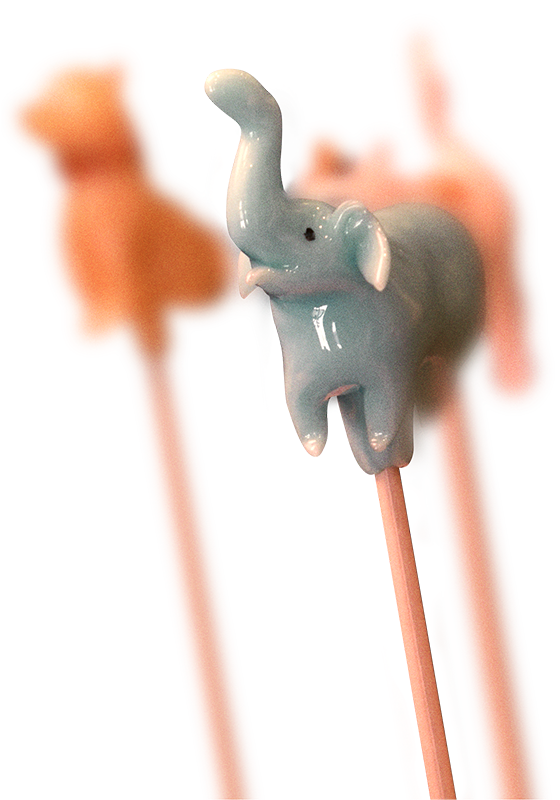 Light blue ceramic elephant figurine perched on wooden chopsticks with its trunk raised upward, photographed against a white background with blurred orange shapes in the distance