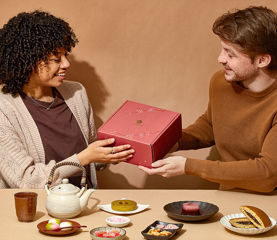 A person is handing a Sakuraco box to another person. With Japanese snacks laid out on a table in front of them.